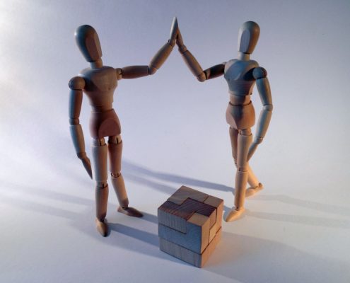 Two artist's mannequins high-five after correctly putting together a wooden cube packing puzzle.